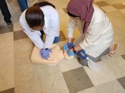 Two women performing CPR on a training manikin in a classroom setting.